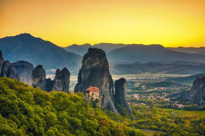 Aerial view of the Meteora rock pillars and the Monastery of Rousanou in Greece at sunset. A forest-covered slope leads to a massive rock formation with a detailed building and tile roof. The sky is a warm golden gradient above a valley and mountains.