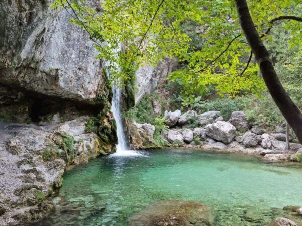 A breathtaking waterfall cascading into a clear turquoise natural pool at Orlias Stream, surrounded by rocks and lush greenery on Mount Olympus, Greece.