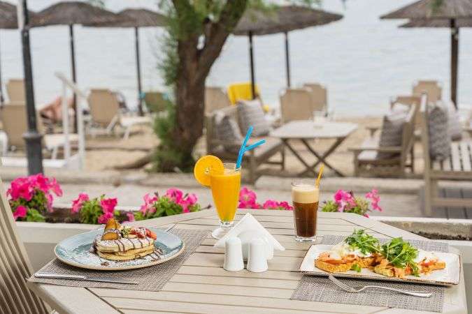 Breakfast table on the Peraia promenade, featuring pancakes, fresh orange juice, iced coffee, and flowers with sea views at Golden Star City Resort, Greece.