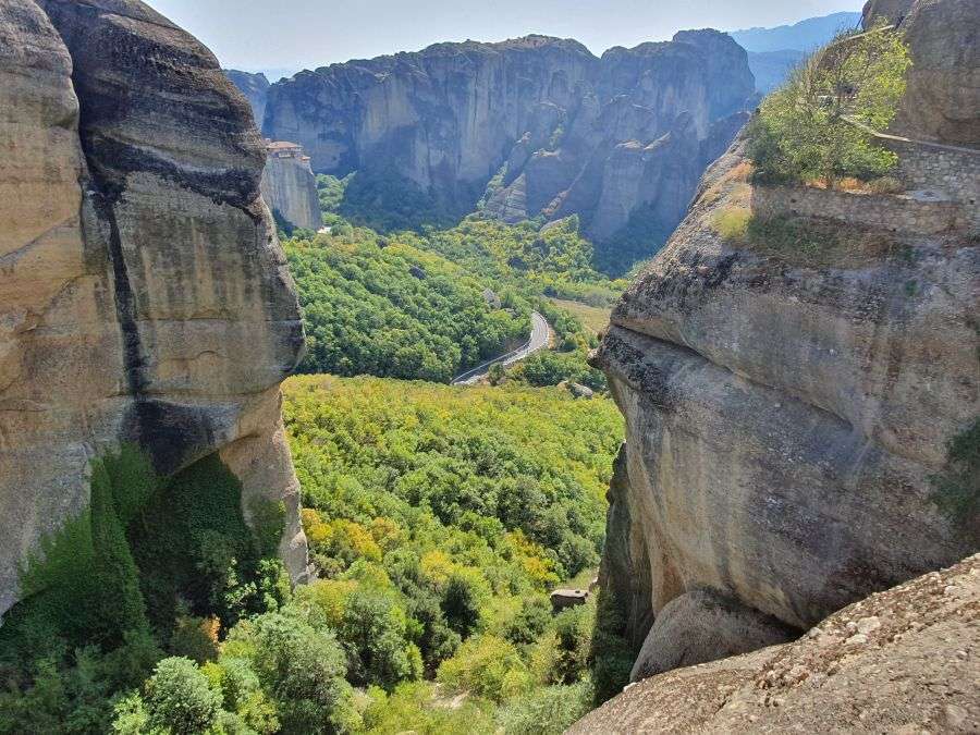 A landscape view of the lush Meteora valley and distant Varlaam monastery, framed by two massive vertical rock cliffs in Greece.